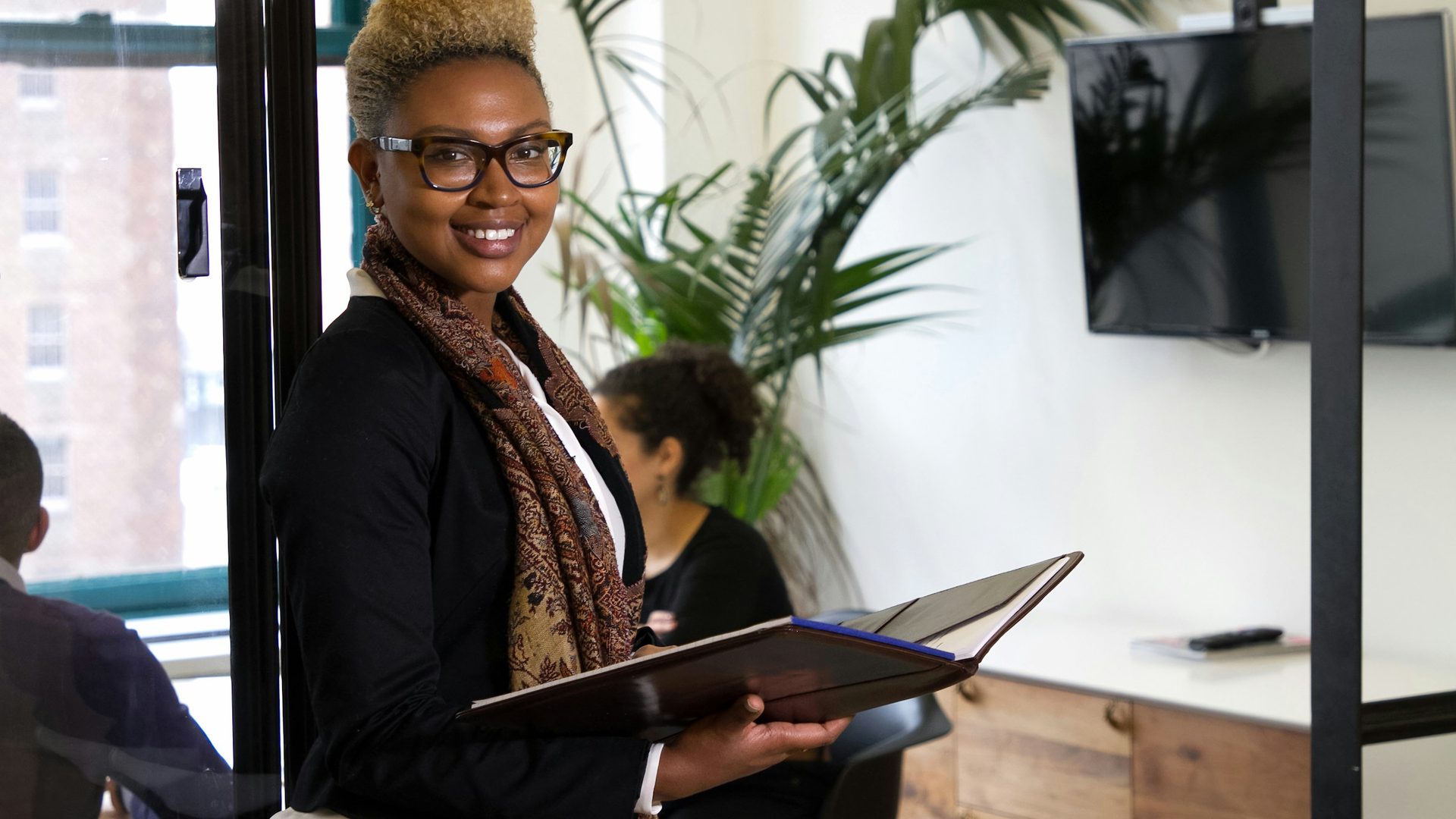 Una professionista con occhiali e foulard sorride in un ufficio luminoso tenendo aperta una cartella di documenti, mentre alle sue spalle i colleghi lavorano